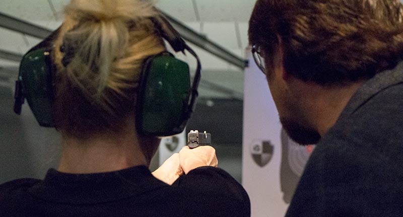 In this photo, firearms trainer and deputy sheriff Paul Carlson teaches a woman how to shoot a handgun for self-defense. The gun selected by the girl is a 9mm Glock 43. They are at a shooting range. A  shooting range, firing range, gun range or shooting ground is a specialized facility, venue, or field designed specifically for firearm usage qualifications, training, practice, or competitions.