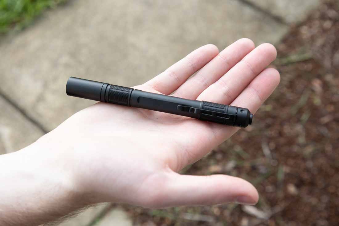 A close-up view of a person holding the Princeton Tec Alloy-X flashlight in their hand, emphasizing the flashlight’s slim and compact penlight shape. The black aluminum body is visible with its integrated steel pocket clip. The tail switch can be seen at the rear, designed for momentary or constant-on operation. The flashlight’s size is noticeably smaller than typical tactical lights, making it suitable for everyday carry in professional or casual clothing. This image illustrates the Alloy-X’s lightweight build and ergonomic grip. It is ideal for commuters, technicians, and anyone needing a rugged but discreet lighting tool. The photo reinforces the flashlight’s low-profile design while still showcasing its tactical features. Princeton Tec Alloy-X flashlight review 