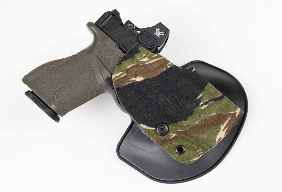 The image shows a Safariland Gravity outside-the-waistband paddle holster with a Springfield Armory Echelon 4.0C pistol seated inside. The holster features a low-cut, open-top design that clears the pistol’s mounted optic without obstruction. Its SafariLaminate shell retains its rigid shape and is wrapped in a Nylon Cordura finish for added durability. The trigger guard of the pistol is completely covered by the holster, preventing accidental contact. The paddle attachment is designed to anchor securely against the body, reducing movement during activity. This particular model is shown in use with the Echelon 4.0C, illustrating the precise molding and audible retention click when the firearm is holstered. The holster’s cut and fit make it suitable for range use, field carry, or concealed carry with a cover garment.