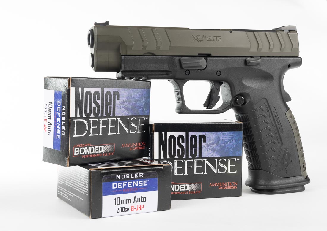 A Springfield Armory XD-M Elite OSP 10mm pistol with a Magpul OD Green Cerakote slide is pictured on a table with Nosler ammunition. The pistol has a polymer frame, forged steel slide, and factory-installed fiber optic front sight. Beside the firearm are Nosler 180 grain and 200 grain jacketed hollow point cartridges, showing the ammunition used in the review testing. The handgun’s optic-ready slide cut and tactical rack rear sight are visible, highlighting its adaptability for red dot optics. The Cerakote green slide provides a durable protective finish that resists holster wear. The pistol’s 16-round magazine design makes it capable for both hunting and self-defense. This image emphasizes reliability across different ammunition types while showcasing Springfield’s finish options.
