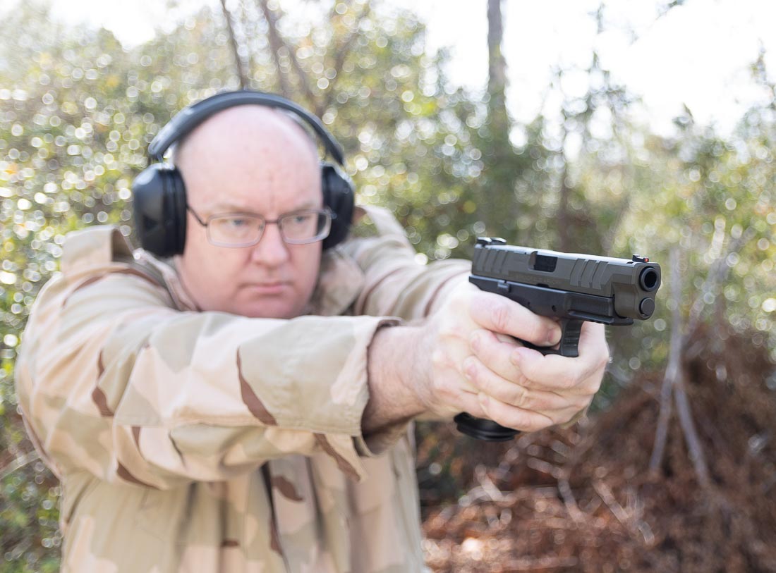 A shooter fires the Springfield Armory XD-M Elite OSP pistol chambered in 10mm Auto at an outdoor range. The handgun has a polymer frame and a Cerakote finish with a factory-installed fiber optic front sight and tactical rack U-notch rear sight. The photo shows the pistol in action, illustrating recoil control and stability in the shooter’s grip. This image highlights the full-size 4.5-inch model, which is optic-ready for red dot installation. The shooter demonstrates a firm two-handed hold, emphasizing the pistol’s ergonomic grip texture and interchangeable backstraps. Visible muzzle rise is modest, showing the manageable recoil of the 10mm cartridge in this platform. The photo reinforces the review’s discussion of the XD-M Elite’s balance, reliability, and practical use as a defensive or hunting handgun.