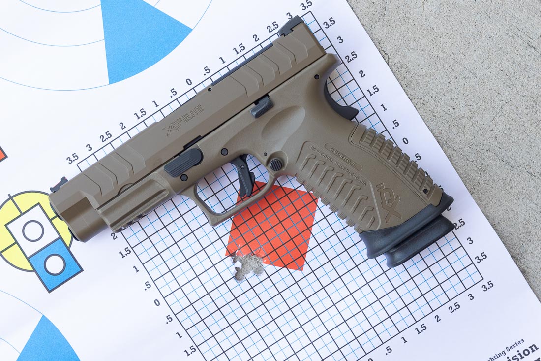 A Springfield Armory XD-M Elite OSP 10mm pistol in Desert FDE finish is placed next to a paper target with tightly grouped bullet holes. The pistol features a polymer frame, Cerakote-coated slide, and a 4.5-inch match-grade barrel. The fiber optic front sight and tactical U-notch rear sight are visible, emphasizing its factory sight system. The target shows a small, concentrated group near the center, confirming the review’s comments on accuracy. This result illustrates the pistol’s ability to produce consistent precision with both practice and defensive 10mm loads. The FDE finish adds durability and visual distinction while maintaining identical performance to the black and green models. This image underscores the combination of accuracy, reliability, and finish options available in the XD-M Elite line.