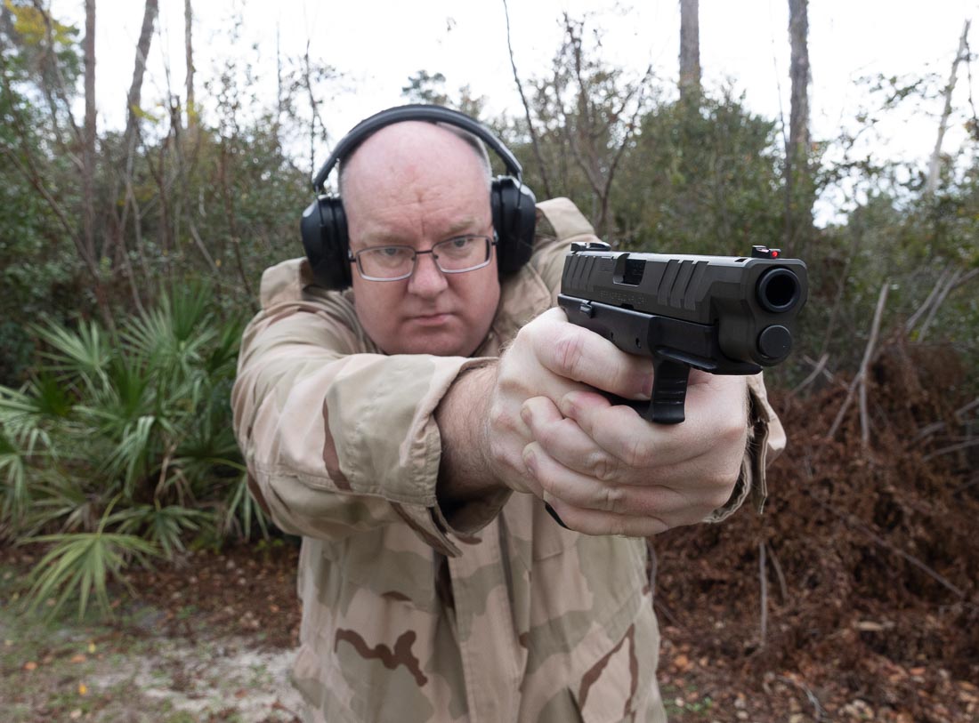 The author is pictured firing the Sportsman’s Warehouse exclusive Springfield XD-M Elite OSP 10mm pistol at an outdoor shooting range. This model has a Magpul OD Green Cerakote slide paired with a standard black polymer frame. The optic-ready slide cut is visible, along with the factory fiber optic front sight and tactical U-notch rear sight. The shooter maintains a two-handed grip, illustrating control over the 10mm cartridge during live fire. Brass is captured mid-ejection, highlighting the pistol’s reliability and smooth cycling. The image demonstrates that this exclusive finish variant performs the same as the standard black and Desert FDE versions. It emphasizes both the cosmetic distinction and the shared performance features across the XD-M Elite 10mm lineup.