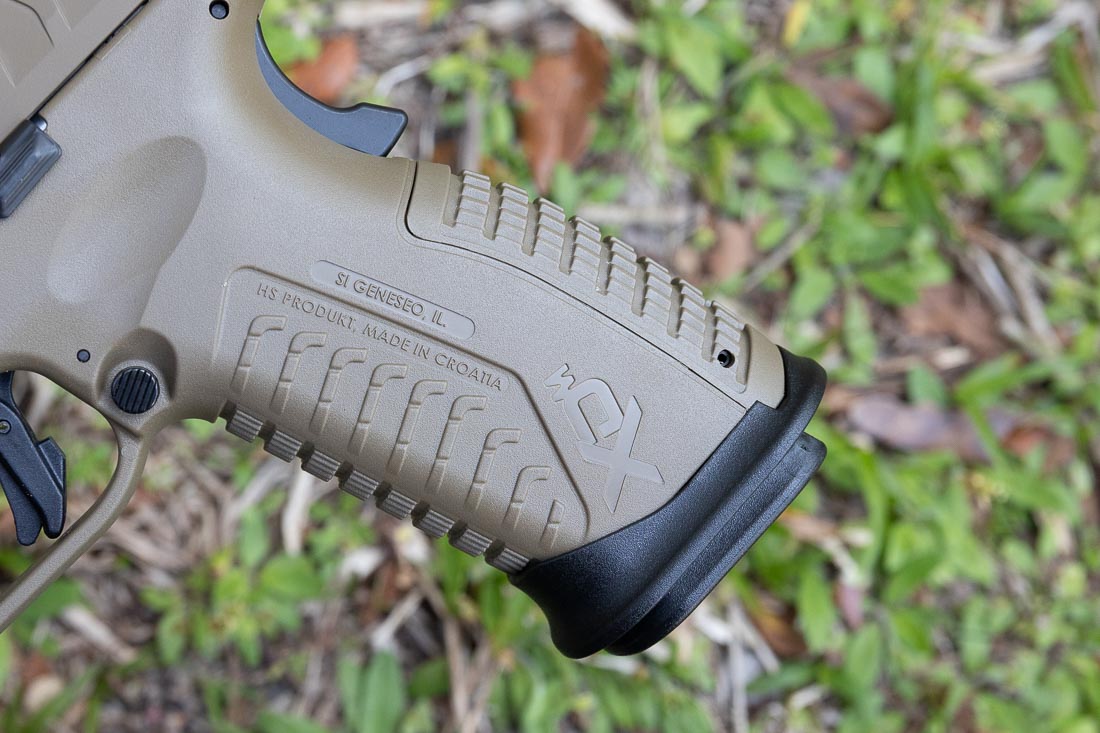 A close-up image of the Springfield Armory XD-M Elite OSP 10mm pistol in Desert FDE finish shows the grip texture and grip safety. The polymer frame has aggressive but comfortable stippling across the front, sides, and backstrap for improved control. The grip safety is clearly visible at the rear of the frame, featuring a raised bump at the base to ensure consistent engagement during firing. This design allows shooters to maintain control even in wet, sweaty, or gloved conditions. The interchangeable backstrap system is also part of the grip, letting users adjust the fit for different hand sizes. The safety system is fully integrated, adding an extra layer of protection against unintentional discharges. This image emphasizes Springfield’s focus on ergonomics, reliability, and safety in the XD-M Elite series.