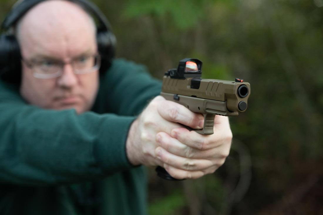 The photo shows noted gun author Richard Johnson firing the Springfield Armory XD-M Elite OSP pistol chambered in 10mm Auto at an outdoor range. He holds the pistol in a firm two-handed grip, demonstrating stability and recoil control during live fire. Brass is seen ejecting cleanly, reflecting the reliability noted in his review. The optic-ready slide, fiber optic front sight, and U-notch rear sight are visible on the handgun. Johnson’s evaluation covered both defensive Nosler ammunition and Blazer Brass practice loads, all of which cycled reliably. The pistol’s Desert FDE finish is also visible in this shot. This image illustrates the hands-on testing that supports the article’s conclusions about accuracy, ergonomics, and performance.