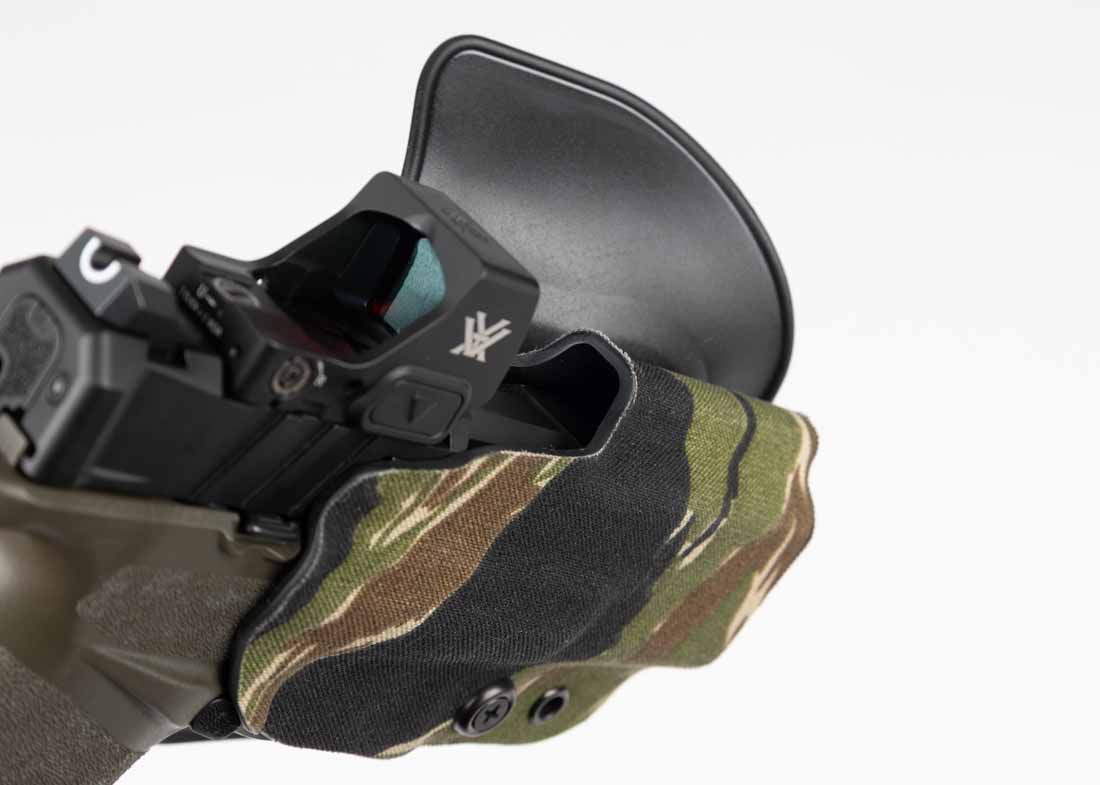 The image shows a detailed close-up of the optics cut on the Safariland Gravity outside-the-waistband paddle holster. The cut is positioned along the upper section of the holster, leaving an open channel around the ejection port and rear deck of the pistol. This space allows clearance for a wide variety of mounted red dot sights, including low-profile optics like the Trijicon RMR and Holosun 507, as well as taller designs such as the Aimpoint ACRO and Trijicon SRO. The edges of the cut are smooth and precisely molded to avoid contact with the optic housing during the draw. This prevents cosmetic damage to the sight and eliminates snag points that could slow access. The design also works with pistols still running iron sights, providing enough front sight clearance for a clean presentation. This compatibility feature makes the Gravity a practical choice for shooters who may switch optics over time.