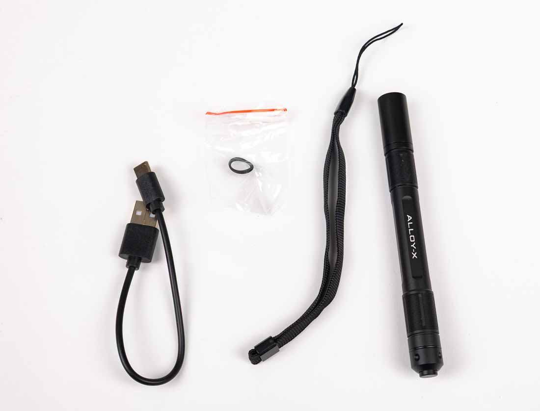 The photo displays all items included with the Princeton Tec Alloy-X flashlight purchase. Shown are the slim black anodized aluminum flashlight, a rechargeable 10900 Li-ion battery with built-in USB-C charging port, and a USB-C cable. The flashlight features an integrated steel pocket clip and tail-mounted switch. The rechargeable battery provides up to 400 lumens output and can be replaced with two standard AAA batteries for backup use. This dual-fuel capability makes the light versatile for both travel and emergency situations. All components are arranged neatly, highlighting the compact nature of the kit. The image reinforces that no external charger or extra accessories are required to start using the Alloy-X immediately.