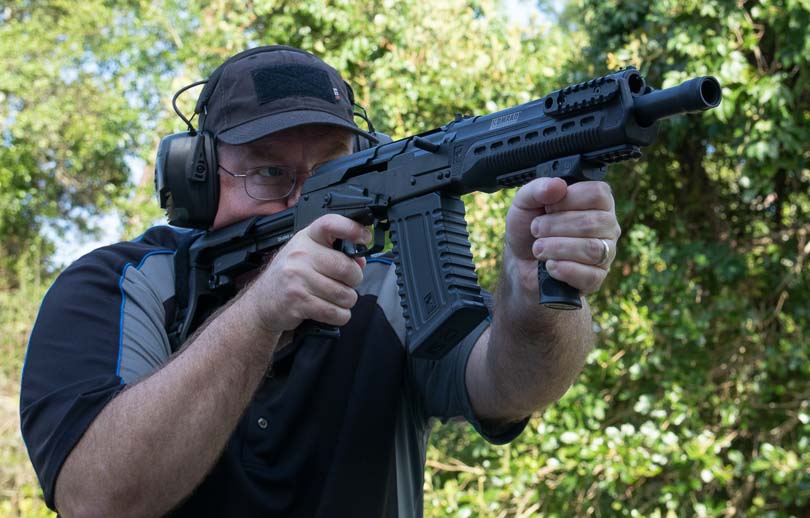The author fires the Kalashnikov Komrad 12-gauge firearm at an outdoor shooting range, demonstrating the compact weapon's handling characteristics with proper shooting stance and arm brace deployment. The Kalashnikov Komrad review highlights semi-automatic performance. Komrad 12 shotgun evaluation tests magazine-fed reliability. AK-style firearms offer familiar manual of arms. Compact 12-gauge platforms deliver defensive capability. Box-magazine shotguns enable rapid reloading potential.