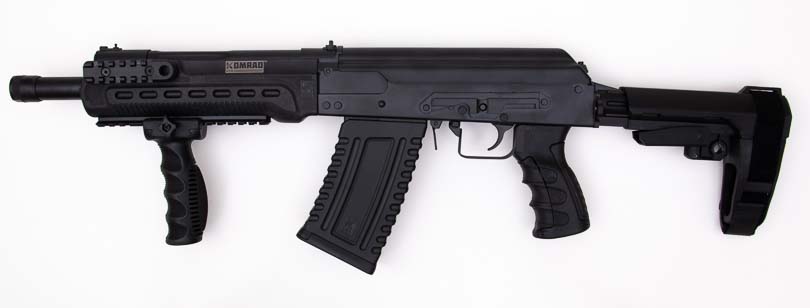 A left-side profile view displays the Kalashnikov Komrad's compact design, showing the 12.5-inch barrel, adjustable arm brace, pistol grip, five-round magazine, and AK-style controls in matte black finish. The Kalashnikov Komrad review examines overall construction quality. Komrad 12 shotgun evaluation highlights gas piston system. Semi-automatic 12-gauge firearms utilize proven mechanisms. Box-magazine shotguns feature AK-pattern ergonomics. Compact defensive platforms measure 31.5 inches overall.