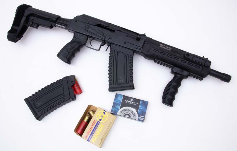 The Kalashnikov Komrad rests on a surface alongside two five-round detachable magazines and various 12-gauge ammunition types, illustrating the complete firearm package and compatible shell options for testing. The Kalashnikov Komrad review evaluates ammunition compatibility thoroughly. Komrad 12 shotgun evaluation tests buckshot and birdshot loads. Magazine-fed shotguns accept 2.75-inch and 3-inch shells. Box-magazine firearms enable rapid tactical reloads. Detachable shotgun magazines hold five rounds capacity.