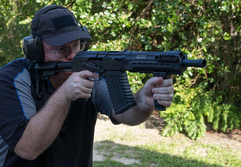 An outdoor range scene captures a firearms reviewer actively testing the Kalashnikov Komrad, a 12.5-inch barreled firearm equipped with an SB Tactical arm brace and vertical foregrip during live-fire evaluation. The Kalashnikov Komrad review examines gas system adjustability. Komrad 12 shotgun evaluation measures pattern performance. Semi-automatic shotgun designs provide quick follow-up shots. Russian-inspired firearms utilize proven operating systems. Detachable magazine shotguns modernize traditional platforms.