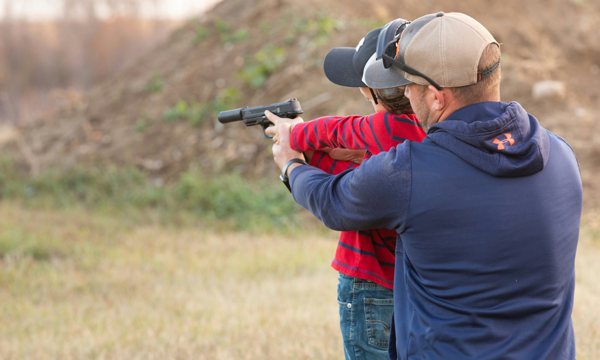 a man helping a woman aim a pistol with a silencer at an outdoor range