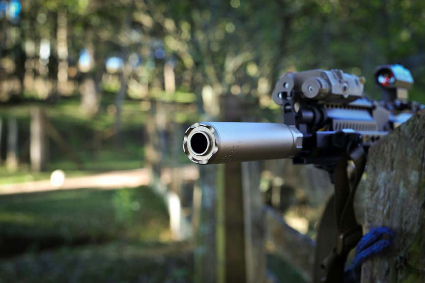 A rifle with a suppressor mounted on a fence, with a blurred outdoor background.
